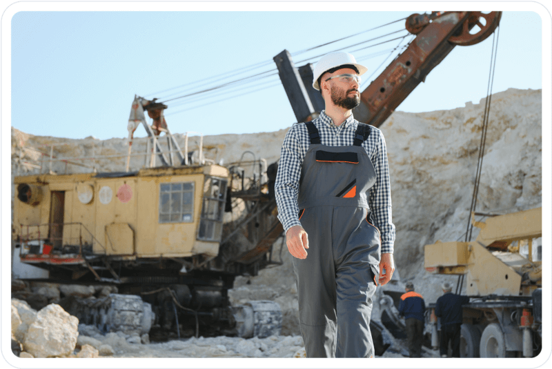 Engineer in safety gear walking through an active mining site with heavy machinery in the background.